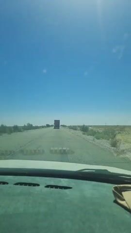 A vehicle drives down a straight, paved road under a clear blue sky. The sun is visible in the upper left quadrant of the frame, casting a glare. Sparse, dry vegetation lines the sides of the road. In the distance, a red truck is traveling in the same direction as the vehicle filming. The view is from inside a vehicle, looking out through the windshield. The dashboard is partially visible in the foreground.