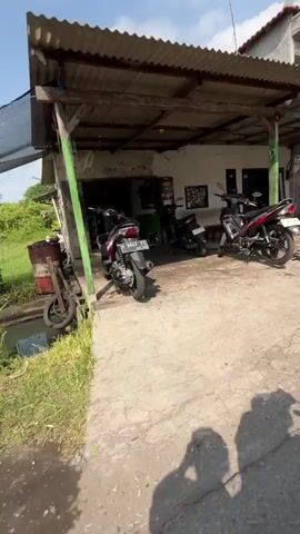 The video shows a simple open-air workshop, likely for motorcycle repair, with a corrugated metal roof supported by wooden and metal posts. Three motorcycles are parked in front of the workshop: a silver scooter on the left, a black motorcycle in the center, and a red and black motorcycle on the right. The workshop interior is dimly lit, with tools and equipment visible. To the left of the workshop, there is a grassy area with a large red drum and a discarded tire. The ground in front of the workshop is paved, and shadows of people are cast across it, indicating the presence of at least two individuals filming the scene. The sky is visible and appears to be clear.