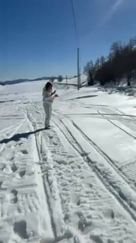 The video shows a person throwing snow into the air on a snowy landscape on a sunny day. The person is wearing a white snowsuit and is standing on a track-like area in the snow. The ground is covered in snow, and there are tracks and footprints visible. In the background, there are trees and a distant mountain range under a bright blue sky. A hand reaches into the frame at the beginning, possibly belonging to the person filming. The audio includes human speech in a foreign language.