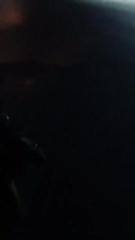 The video begins with a dark frame that quickly transitions to reveal a nighttime scene. The camera appears to be mounted on a moving vehicle, possibly a motorcycle, indicated by the blurry foreground showing part of the vehicle. The vehicle is traveling on a paved road. In the background, there are buildings and a green truck parked. Streetlights illuminate parts of the scene. The camera pans slightly as the vehicle moves forward, offering a view of the road ahead, the buildings to the side, and the parked truck. The overall lighting is dim, characteristic of a nighttime environment. There are no people visible in the frame.