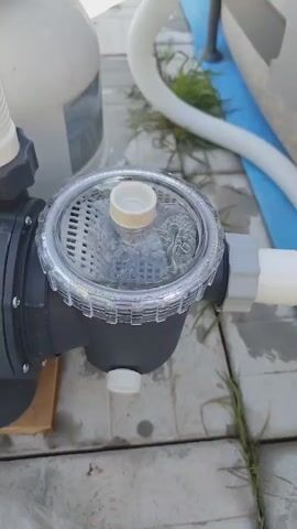 The video shows a close-up of a pool pump's strainer basket, which is filled with water and some debris. The strainer basket is made of clear plastic, allowing visibility into the basket. Inside, a white mesh filter is visible, and small bubbles are rising to the surface of the water, indicating the pump is running. A white cap sits on top of the filter. The pump itself is dark gray and has white fittings. The strainer basket is connected to a larger, white plastic component, possibly a filter tank, in the background. A white hose snakes away from the pump to the right. The pump and its components are situated outdoors, on a paved surface, likely a patio or deck. There are patches of dry grass growing through the paving stones, suggesting an outdoor, possibly unkempt, setting. A portion of what appears to be the edge of an inflatable pool is visible in the upper right corner of the frame, colored blue. The lighting suggests it is daytime, with no specific indication of the time of day or weather conditions. No people are visible in the frame. There is no discernible text in the video. The overall scene is that of a functional outdoor swimming pool setup, focusing on the operation of the pool pump.