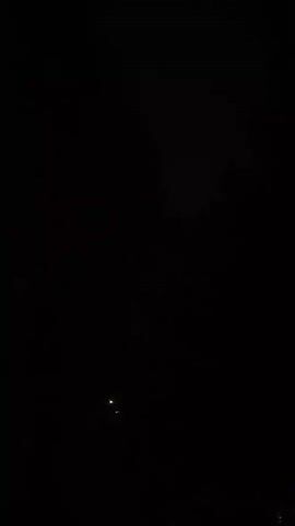 The video shows a brief glimpse of a dark outdoor scene. Two faint, yellow lights are visible in the distance. Above these lights, the dark silhouettes of trees are discernible against a dimly lit sky.