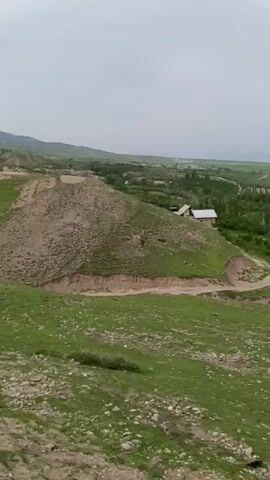 The video presents a landscape view of a hilly terrain with a village in the distance. The foreground consists of grassy slopes with some rocky patches. A dirt road winds through the hills and leads towards the village. In the distance, mountains are visible under a cloudy sky. The village is nestled in the valley, with houses surrounded by trees and fields. The camera pans slowly across the scene, providing a wide view of the landscape.