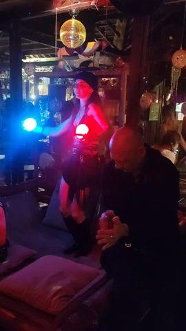 A woman in a dark outfit and knee-high boots performs with red and blue orbs in a dimly lit bar. A man sits on a nearby couch, playing a tambourine. Other patrons sit around the room, and a disco ball hangs from the ceiling.