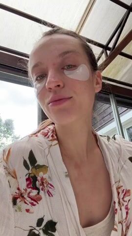 A young woman with fair skin, hazel eyes, and light brown hair pulled into a ponytail is eating a piece of toast topped with what appears to be salmon roe. She is wearing white under-eye patches and a light-colored floral robe over a ribbed tank top. The woman is inside what looks like a sunroom or enclosed porch, as a window with a view of the outside is behind her, showing green trees and foliage. The roof is made of translucent panels. The camera angle is from below, looking up at the woman. She smiles while taking a bite and chewing the toast. The focus remains on the woman and her actions. A male voice is heard making comments, but he is not seen in the frame.