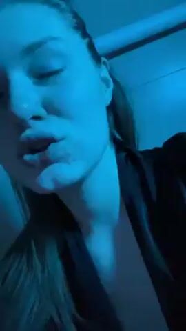 In a dimly lit, close-up video, a young woman with fair skin and light brown hair is seen making exaggerated pursing motions with her lips. Her hair is pulled back from her face. She is wearing a black, v-neck garment. The lighting is predominantly blue. In the background, a white, slightly curved object that appears to be a pipe is visible.