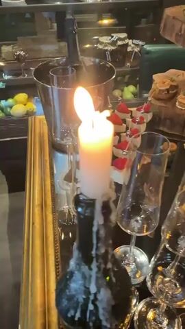 The video shows a buffet display with a variety of appetizers and champagne glasses. There are four types of appetizers, including ones with cheese and lemon, shrimp, beets, and strawberries. The appetizers are arranged on a black table and a wooden tiered stand resembling a tree trunk. There's a metal ice bucket with a bottle of champagne, and a dark glass bottle used as a candle holder with a lit white candle. The background shows a glass display case with various desserts and fruits.