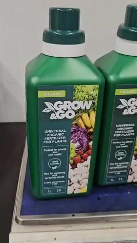 The video shows three green plastic bottles of fertilizer placed on a scale. The bottles have white caps and green labels with the brand name "GROW & GO" printed on them. The labels also indicate that the product is "ORGANIC UNIVERSAL ORGANIC FERTILIZER FOR PLANTS" and "ORGANO-MINERAL COMPLEX LIQUID FERTILIZER FOR BERRIES AND VEGETABLES". The bottles are filled with a liquid and are in a setting that appears to be a store or warehouse. The video is a close-up shot of the bottles, with the focus on the labels.