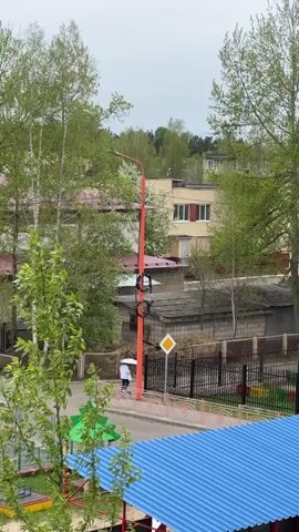 The video shows an outdoor scene in a residential area. In the foreground, there's a colorful structure, possibly a children's play area, with a blue roof and green and red accents. A pedestrian crossing sign is also visible nearby. An orange street lamp stands tall beside the road. Beyond the play area and the road, there's a fence separating the area from some garages. A yellow building with red accents on the windows is visible behind the garages. The scene is interspersed with trees, their leaves indicating it's likely spring. A car pulls up to the right and parks. The sky is overcast, suggesting a cloudy day. The video appears to be taken from an elevated position, such as an apartment building.