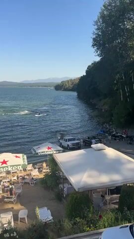 The video shows a scenic waterfront view on a sunny day. The foreground features a beach area with lounge chairs and umbrellas, some of which are branded with the Heineken logo. People are seen relaxing on the beach and under the umbrellas. Adjacent to the beach, there is a paved area with several vehicles, including white and black pickup trucks, and what appears to be a jet ski. The water is calm with a few boats and a person on a jet ski actively riding on the lake. The background reveals a lush, tree-covered hillside sloping down to the water, and distant mountains under a clear blue sky. The overall impression is a vibrant, recreational atmosphere at a lakeside resort or beach club.
