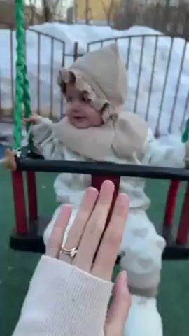 A video shows a baby on a swing. The baby is wearing a beige outfit and hat, with white boots or booties, and is secured in the swing seat with a harness. The swing is on a playground with a green surface, and patches of snow are visible in the background. A person's hand, wearing a ring, pushes the swing gently. The person also says "up" and later "поймаю, поймаю, поймаю" (pomayu, pomayu, pomayu), which translates to "I'll catch you, I'll catch you, I'll catch you" in Russian.