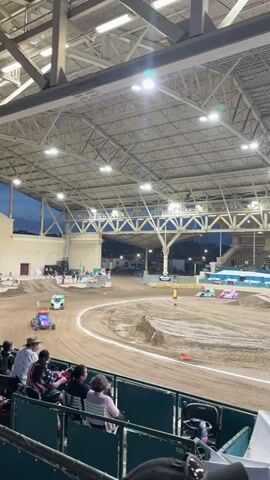 The video shows an indoor dirt track with several remote-controlled cars racing. The track is made of dirt and has several ramps and turns. There are spectators in the stands watching the race. The overall lighting suggests it is either early evening or indoors with artificial lighting. The cars are small and colorful, and they appear to be moving at high speeds. There are a few people standing near the track, possibly working on the cars or officiating the race. The structure of the building appears to be a large arena with a metal roof and exposed beams.