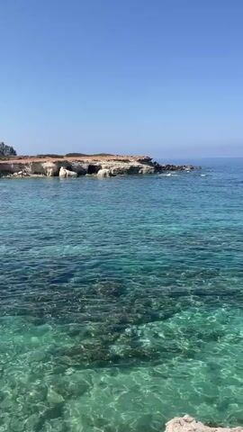 The video shows a scenic view of a rocky coastline and the ocean. The water is a clear turquoise color, and the seabed is visible in the shallow areas. The sky is a bright blue, with a few wispy clouds in the distance. On the right side of the frame, a shipwreck is visible, partially submerged in the water. The shipwreck appears to be an old cargo ship, with its rusted hull and broken masts visible. The waves are gently lapping against the shore, and the sound of the ocean can be heard in the background. The overall impression is one of peace and tranquility, with a touch of melancholy due to the presence of the shipwreck.