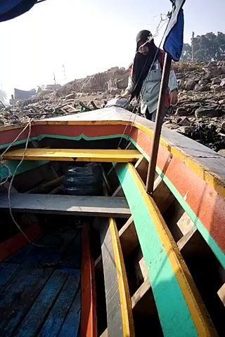 A person wearing a hijab and a light-colored jacket over a pink shirt, along with dark pants and boots, is in a boat. The person is positioned near the front of the boat, kneeling and reaching out. The boat appears to be made of wood with colorful painted accents in red, orange, yellow, and green. Inside the boat, there are wooden benches and a black barrel. The background shows a rocky shore with some structures and vegetation under a bright sky. The person is looking towards the camera and smiling. The camera perspective is from inside the boat, looking towards the front and up at the person. The video captures a series of movements as the person adjusts their position and interacts with the boat.