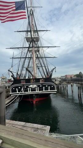 This video shows a rear view of a historic ship docked at a pier. The ship, the USS Constitution, is a three-masted frigate. It is painted black with a red hull and has an American flag flying from its stern. The ship is docked alongside a wooden pier, and the water around it is dark green. The sky is cloudy and overcast. The video is shot from a slightly low angle, looking up at the ship. The camera is stationary.