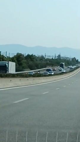 A daytime aerial view shows a highway with multiple lanes of traffic. On the left side of the road, a line of vehicles is stopped or moving slowly. These vehicles include cars and at least one large truck. The highway curves to the right, with more cars visible in the distance. The background consists of a treeline, power lines, and distant hills under a hazy sky. The perspective suggests the viewer is looking down from a slight elevation.