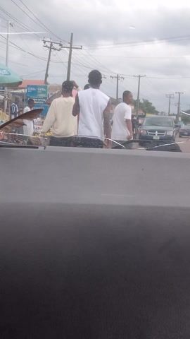 This video captures a street scene, likely in Aketu-Oja, Nigeria, under an overcast sky. The footage appears to be filmed from inside a vehicle, looking out onto a road with several people and objects.

The primary subjects are a group of men standing on the side of the road, some wearing white shirts and one in a cream-colored outfit. Their postures suggest they might be waiting or observing something. Further down the road, a vehicle is visible, with its headlights on, indicating it might be dusk or a particularly cloudy day.

To the right of the road, there's a structure with a green awning and a metal fence. A woman in a blue and patterned dress and a white head covering is standing next to a motorcycle. The motorcycle is a prominent object in this part of the scene, with visible wheels and frame. Behind her, there are various items covered in black plastic, possibly goods or supplies.

There are utility poles with wires running along the side of the road, a common sight in many urban and semi-urban areas. Signs are partially visible, including one that reads "DSTV" in blue and green letters, suggesting the presence of a telecommunications service provider. Another sign has the words "NOW OFFICE" and "LOKAL".

The overall atmosphere seems ordinary, a snapshot of everyday life on a Nigerian street. There are no obvious signs of distress or celebration, but rather a sense of calm or routine activity. The cloudy weather contributes to a diffused light, making it difficult to pinpoint the exact time of day, though the vehicle headlights suggest it's not the middle of the afternoon.