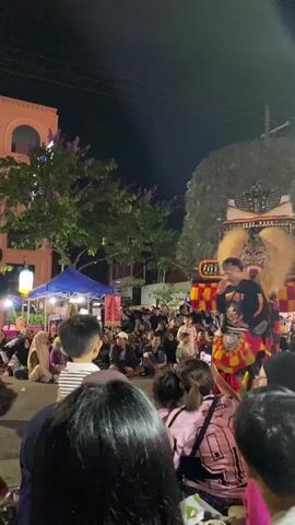 This video captures a lively street performance at night, likely in Madiun, Indonesia, given the context. The central focus is a performer dressed in an elaborate, traditional costume featuring a large, fierce mask with a prominent mane and what appears to be a headdress made of dark feathers. The costume is vibrant, with red, yellow, and black fabrics.

The performer is being assisted by another individual, who is dressed in dark clothing with a white belt and also has some colorful fabric attached to their legs. They are walking among a crowd of spectators who are gathered on the street, watching the performance. Many of the onlookers are seated on the ground or standing, and several are holding up their phones to record the event, indicating a sense of excitement and engagement.

In the background, there are buildings with warm lighting, trees, and tents, suggesting a festive atmosphere, possibly a fair or cultural festival. A street lamp is visible, casting a soft glow, and the sky is dark, confirming it is nighttime. The overall mood appears to be joyful and celebratory.

There is some visible text on the performer's shirt and on a banner or sign in the background. The shirt has a logo that is difficult to decipher clearly, but it appears to be text-based. The banner in the background is also partially obscured and the text is not legible. The prominent mask with its elaborate details and the engaged crowd are the most striking elements of the scene.