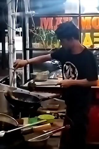 A man in a black t-shirt is cooking at an outdoor food stall. He uses a ladle and a pair of metal tongs to stir food in a wok over a flame. Various cooking utensils sit on the counter next to the wok. Behind the cook is a lighted sign with the words "MAX MO TEA" on it.