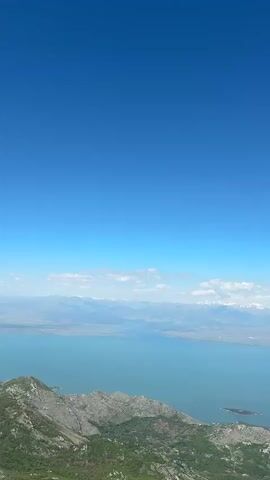 The video presents a wide shot of a mountain landscape, taken on a clear, sunny day. In the foreground, rugged, green mountains rise, speckled with vegetation.  Beyond the mountains lies a large body of water, possibly a lake, with a turquoise hue. In the distance, snow-capped mountain peaks fade into the horizon, partially obscured by light clouds. The sky above is a vibrant, solid blue. A small contrail appears briefly in the upper center.