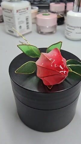 The video displays a close-up of a decorative red rose with green leaves, crafted from delicate materials, resting on a black, cylindrical container. The rose appears to be made of translucent, shiny material, possibly resin or plastic, with gold wire accents outlining its petals and leaves. The leaves are a translucent green, also with gold wire detailing. The black container has a matte finish and a slightly textured surface.

In the blurred background, several jars and containers are visible on a white surface. Some of these containers appear to be cosmetics or skincare products, judging by their packaging, with labels indicating potential brands. One white box-like container in the upper left has prominent text that appears to be in English, possibly related to a product. Other jars are round and colored pink and white, with some having visible lids. A silver-capped bottle is also visible. The overall setting seems to be a crafting or personal care area, possibly a desk or countertop.

The lighting suggests an indoor environment, with soft lighting that highlights the glossy texture of the rose. The focus is on the rose and the container, with the background softly blurred, creating a shallow depth of field. No people are visible in the video, and the activity taking place is implied to be the placement or display of this decorative item. The atmosphere is still and focused on the artistry of the rose. There are no overt clues about the time of day, weather, or specific emotions, other than a sense of quiet craftsmanship. The location context of Abinsk, Russia, is not visually represented in the footage.