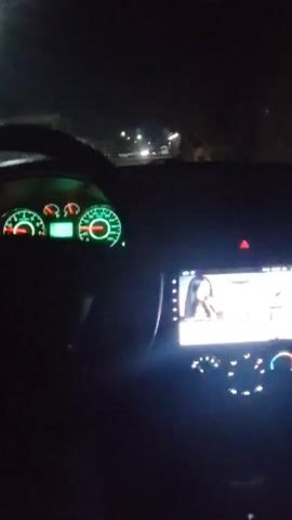 The video shows a driver's view from inside a car at night. The dashboard is visible, with illuminated green gauges. A screen on the center console displays a video of a woman with long dark hair and dark clothing. The car is moving along a street lined with buildings and other parked cars, illuminated by streetlights.