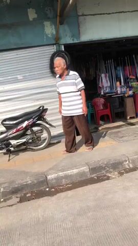 An elderly man with gray hair walks along a sidewalk. He is wearing a striped shirt and brown pants. A motorcycle is parked on the sidewalk to his left. Behind him, there is a store with various items displayed outside. The scene appears to be outdoors, with sunlight casting shadows.