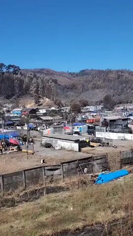 The video shows a small town with many of the structures covered by blue tarps. The hills behind the town are scarred by fire, with many trees reduced to blackened stumps. There is a low concrete wall along the bottom portion of the frame.