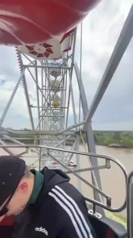 The video shows a Ferris wheel ride with two passengers in the car. One person wears a black hat, sunglasses, and an Adidas jacket. The other passenger is a blonde woman. The camera angle is from inside the car, looking out at the wheel’s structure, other cars, and the view from the ride. The view reveals a wide river with trees lining its banks and glimpses of buildings. As the ride ascends, the video captures the amusement park layout below, including rides like a rollercoaster and structures like a red-and-white tower. The sky is cloudy.