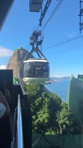 The video captures a modern cable car gondola suspended against a vibrant blue sky, in what appears to be a station or terminal. The white, oval-shaped gondola has large, clear windows through which several passengers are visible, some standing and observing the view. It is connected to thick cables overhead by a sturdy metal support structure.

In the immediate foreground, to the lower left, part of a metal railing and a person's shoulder suggest the viewer is on a platform. Below the gondola, lush green foliage covers a steep hillside, indicating significant elevation. To the right, a large green structural element, likely part of the cable car station or a support, frames the view.

Dominating the left background is a prominent, rocky mountain, characteristic of Sugarloaf Mountain in Rio de Janeiro, Brazil. Its peak is visible, and its slopes are covered in patches of green vegetation. Beyond the immediate landscape, a vast expanse of sparkling blue ocean or bay stretches into the distance, dotted with other distant mountains and coastal features, typical of the Rio de Janeiro coastline.

The gondola is stationary, possibly having just arrived or waiting to depart, allowing passengers to take in the panoramic scenery. The weather is clear and sunny, with a bright blue sky and no clouds, suggesting a perfect day for aerial sightseeing. The lighting indicates it is likely mid-day. No discernible text is visible on the gondola or prominent structures. The overall scene evokes a sense of travel, sightseeing, and breathtaking natural beauty.