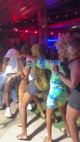 A group of young women are dancing in a dimly lit outdoor venue. Red and blue lights are illuminating the scene from above. The women are mostly barefoot and wearing casual clothing like shorts and t-shirts. One woman in the foreground is wearing a colorful, patterned dress. They are all facing away from the camera and appear to be moving in unison to music, with some of them raising their hands and pointing. In the background, a mural is visible on a wall.