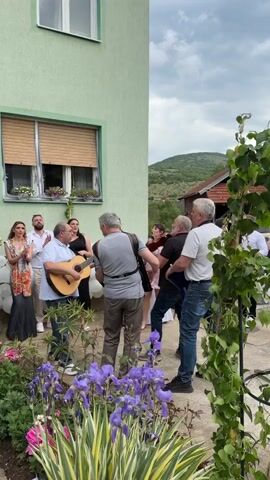 A group of about ten people is gathered outside of a green house, enjoying live music. A man plays a guitar, another plays a violin, and someone else seems to be playing the accordion. Some people are clapping, and others are singing or dancing. The house has a window with flower boxes, and there are balloons nearby, possibly indicating a celebration. The landscape includes flowers in the foreground and hills in the distance.