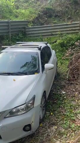 A white car is parked on a gravelly, overgrown roadside. The car is oriented facing left, with its rear end visible. To the right of the car, a large pile of branches and debris is stacked haphazardly. Beyond the debris, a narrow, rocky stream flows. The stream is surrounded by lush green foliage and trees, suggesting a natural, wooded environment. The water in the stream appears to be moving rapidly, with some small rapids visible. The camera pans from the car and debris towards the flowing water and the surrounding greenery.