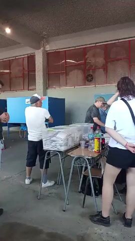 The video shows an indoor scene at a polling station. Several people are visible. A man in a white t-shirt and dark shorts stands next to a blue voting booth with the number 477 on it. There is a table with chairs around it, covered with stacks of papers in clear plastic bags. Drinks and cups are also on the table. Two men are positioned near the table, and a woman in a white shirt and black shorts stands with her back to the camera. Another person is near the table attending to the papers. The setting appears to be a school or gymnasium type building with large windows behind the people at the table.