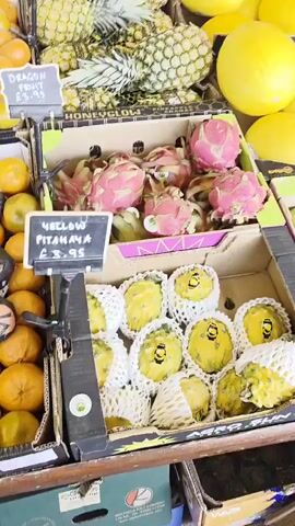 The video shows a display of various fruits for sale at what appears to be an outdoor market or store. The fruits are arranged in cardboard boxes and include pineapples, yellow dragon fruit, red dragon fruit, yellow melons, mandarins, kiwis, apples, and a cantaloupe melon. There are price tags displayed for some of the fruits. The scene suggests a focus on fresh produce. The camera appears to be held by someone walking past the fruit display.