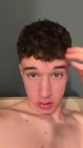 A close-up shot features a young man with short, curly brown hair and green eyes. He is fair-skinned and appears to be shirtless, with part of a light-colored bed frame visible in the background. The man is looking directly at the camera and speaking. He gestures with his right hand, touching his hair at the start of the clip. The background is a plain gray wall.
