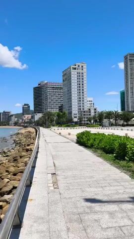 A paved walkway runs alongside a rocky shoreline with the ocean on the left. In the background, several modern high-rise buildings stand against a bright blue sky with scattered white clouds. The camera pans slowly to the left, showing more of the coastline and the ocean stretching towards a distant mountainous horizon.