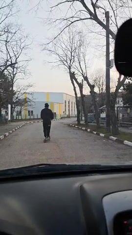 The video, shot from inside a vehicle, shows a person riding an electric scooter on a paved road. The individual is wearing a dark-colored jacket, pants, and a dark blue beanie. The road is lined with a black and white curb, and on the left is a building with white and yellow panels. The environment suggests a residential or industrial area with bare trees and other buildings visible in the background. The sky appears overcast, indicating a cool or potentially cold day. The vehicle seems to be following the scooter, maintaining a consistent distance.