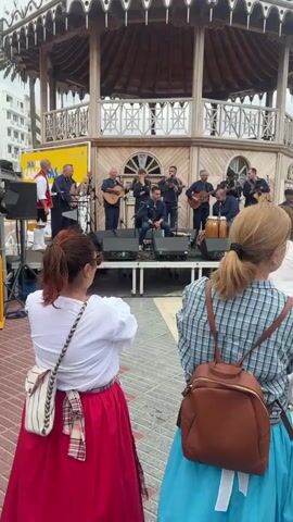 Here is a description of the video:
The scene takes place outdoors on a cloudy day. A band is performing on an elevated, white platform under an ornate gazebo with a dark, multi-tiered roof. The band consists of approximately a dozen men, mostly middle-aged, playing instruments including guitars, percussion, and some wind instruments. One man is seated in the center and appears to be a singer. They are dressed in dark jackets, with one member in a traditional red and white outfit.
In the foreground, two women are facing the band. The woman on the left is wearing a white blouse and a bright red skirt, with a patterned bag slung across her shoulder. She begins to clap her hands. The woman on the right is wearing a plaid shirt and a blue skirt, with a brown backpack. Other people are visible in the periphery of the shot, also watching the band. A banner reading "Día de Canarias" is visible on the left side of the frame. The ground is paved with red and beige brick tiles. In the background, there is a low building, and a palm tree is visible to the left.