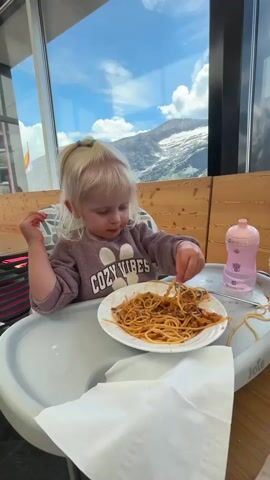 A young child with blonde hair pulled into a ponytail sits in a high chair, eating spaghetti with chopsticks. The child is wearing a purple sweatshirt with "COZY VIBES" written on it. A plate of spaghetti and a pink bottle are on the tray in front of the child. A large window behind the child looks out onto a mountain landscape with a snow-capped peak. The scene appears to be an indoor dining area with wooden paneling. The video is a short loop, showing the child picking up spaghetti and bringing it to their mouth.