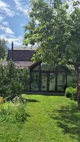 The video shows a green lawn in front of a house with a large glass conservatory. The conservatory has a triangular roof and is surrounded by trees and bushes. Sunlight filters through the leaves of the trees, casting dappled shadows on the grass. A chimney can be seen on the roof of the house.