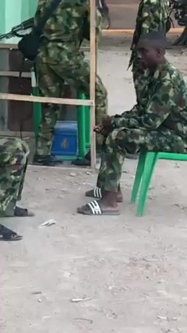 The video shows several people in camouflage uniforms, possibly military personnel, in an outdoor setting. One person is seated on a green plastic chair, wearing Adidas-branded slides. Another person stands nearby, and parts of other people in uniform are visible in the frame. There is a small table with green accents next to them, with a blue object underneath. The ground appears to be a light-colored dirt or sand.