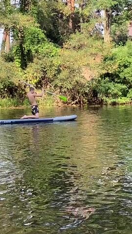 A person kneels on a paddleboard in a body of water, holding a paddle and paddling. The person is wearing swim trunks. Trees and foliage line the background of the water.