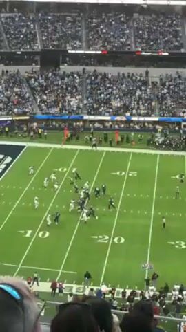 The video shows an American football game in a packed stadium, filmed from an elevated spectator position. The teams, identifiable by their uniforms, are aligned on the field. One team, in dark uniforms, appears to be on offense and passes the ball. The crowd, largely clad in blue, fills the stands, with many people visible watching the game. The field has yard lines marked, along with the name "Chargers" painted in the end zone. The overall environment appears to be a professional sporting event with a large audience.