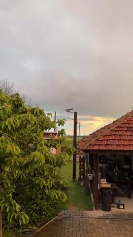 The video shows a static, medium shot of a rural scene under an overcast sky. To the left, a leafy green tree obscures part of the view.  Behind the tree, a glimpse of another house can be seen.  In the foreground to the right is the corner of a structure with a red tiled roof. A paved walkway leads down from the viewpoint.  Beyond the structure, a green field is visible, with utility poles and power lines in the distance.  The sky is a mix of gray clouds and a sliver of pale golden light near the horizon. No people are visible.