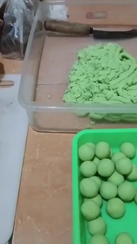 The video shows a close-up shot of a kitchen counter. In the foreground, a bright green plastic tray is filled with small, uniformly shaped green balls. Behind this tray, a clear plastic container holds a pile of light green dough or clay with a textured surface. A wooden-handled knife with a dark blade is placed on top of the container. To the left of the container, a white cutting board is visible. The counter surface is a light brown color with a tiled appearance. The background is slightly out of focus, with a glimpse of a plastic bag and other items. The lighting appears to be artificial, casting a warm glow on the scene. The overall impression is one of food preparation or crafting.