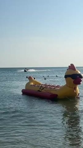 In the video, a person rides a jet ski across a calm, blue body of water under a clear, bright sky. The jet ski is moving at a moderate speed, leaving a white spray in its wake. In the foreground, a large, inflatable yellow and red tube-like object is partially visible, floating on the water. Several people are in the water behind the inflatable object, appearing to be swimming or relaxing. The overall scene depicts a recreational activity on a sunny day at the beach or a similar body of water.