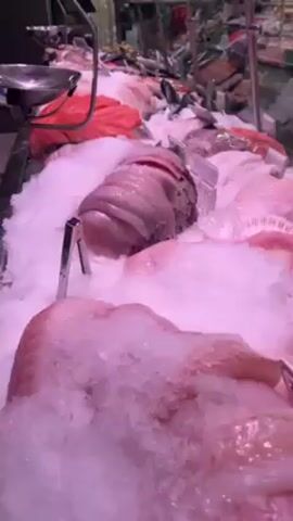 The video shows a fish display in a store or supermarket. The fish are laid out on a bed of crushed ice. Varieties of fish are visible, including whole fish and cut fillets. Some fish are pink, while others are white. Small price tags are placed next to each type of fish. In the background, there are shelves stocked with other food products.
