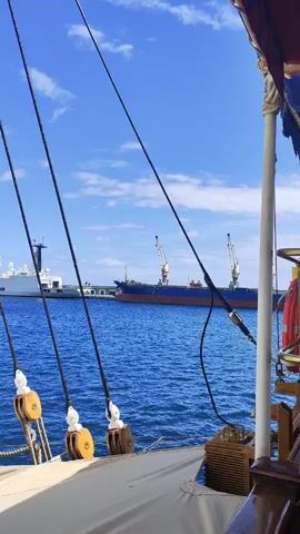The video displays a serene view of a body of water under a bright blue sky with scattered clouds. The foreground shows part of a wooden sailing ship, with ropes and wooden pulleys visible. In the mid-ground, a large blue and red cargo ship is anchored, equipped with two cranes. In the background, a white cruise ship is docked at a pier. The overall atmosphere is peaceful, suggesting a calm day at sea or in a harbor.