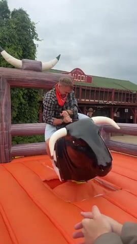 This is a short video clip of a person riding a mechanical bull. The rider, a young man wearing a plaid shirt, light-colored shorts, and pink socks, is positioned on the bull's back, gripping its handles. The mechanical bull is black and white with brown markings, and it's situated on an orange inflatable platform, suggesting an amusement park or event setting.

In the background, there's a wooden structure resembling a saloon, complete with a sign that reads "SALOON" in red letters. The building has a rustic appearance and is situated behind a wooden fence. Lush green trees and foliage are visible to the left of the frame, indicating an outdoor setting. The sky is overcast, suggesting a cloudy day.

The video shows the mechanical bull in motion. In the first second, the rider is leaning forward. In the second second, the bull bucks, and the rider is thrown back, almost falling off. The video then reverts to the initial position. The overall mood appears to be lighthearted and recreational, with the rider likely enjoying the challenge of staying on the bull. The visible text "SALOON" on the building in the background provides a clear location cue for the scene. The timestamp indicates the video was recorded in Mozhaisk, Russia.